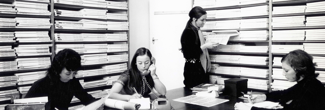 black and white photo of women working at a large desk with lots of paper work stacked behind them