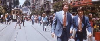 People in the 1970s walking along Bourke Street. Two men in suits are in the foreground. 