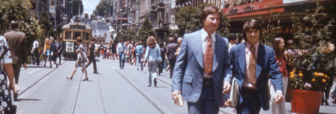 People in the 1970s walking along Bourke Street. Two men in suits are in the foreground. 