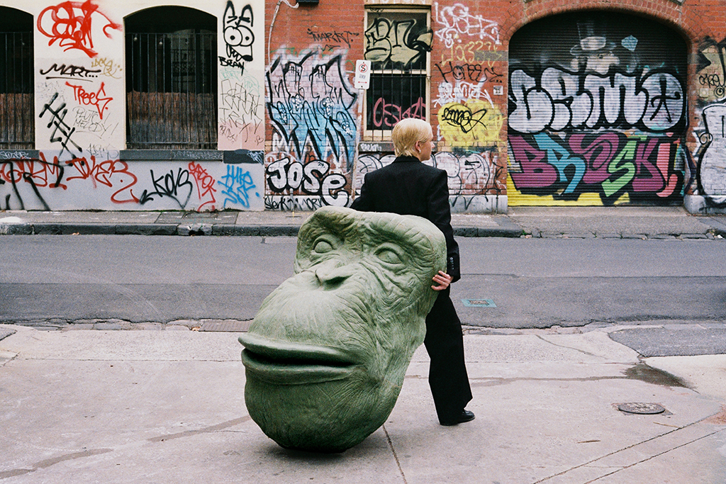 A colour photograph of a person dragging a large prop of a chimpanzee's head through a Melbourne city alleyway