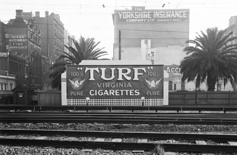 black and white photo of turf billboard along side train tracks with buildings and palm trees in the background