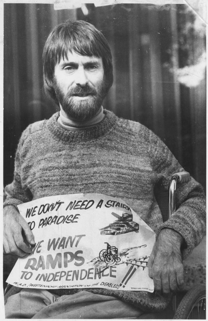 Black and white photograph of Geoff Bell in his wheelchair, holding a sign that says We don't need a staircase to paradise. We want ramps to independence