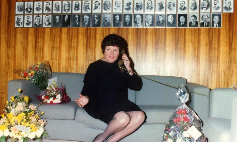 Colour photograph of Joan Kirner sitting on a couch, talking on the telephone, with photographs of past Premiers on the wall behind her