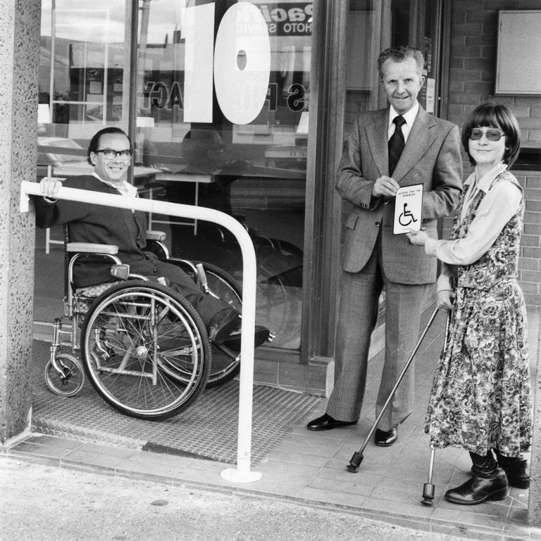 Black and white photograph of three people outside a bank. A man using a wheelchair is on a newly installed ramp.